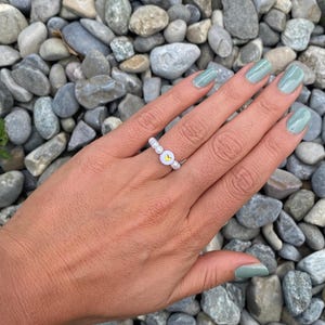 May include: A beaded ring with white beads and a central charm featuring a yellow heart on a white background. The ring is worn on a hand with light green nail polish. The background is a collection of gray stones.