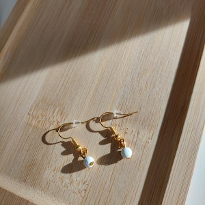 Könnte beinhalten: Ein Paar goldfarbene Ohrringe mit weißen, runden Perlen. Die Ohrringe werden auf einem hellbraunen Holztablett präsentiert, mit Schatten. Die Ohrringe haben ein schlichtes, elegantes Design.