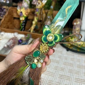 Pode incluir: Uma varinha decorativa com um grande cristal facetado de cor verde-azulado. A varinha é adornada com enfeites florais verdes e dourados, pedras preciosas verdes, pérolas e penas castanhas. Uma borla castanha está presa à base.