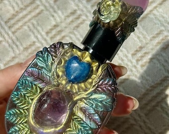 May include: An ornate perfume bottle with a black cap and a decorative design. The bottle features a purple crystal ball on top and a central purple stone, surrounded by iridescent green and blue leaf-like details. A blue heart-shaped stone is also present.