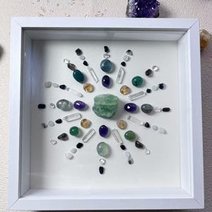 May include: A square picture framed in white, featuring a crystal mandala design. The mandala is composed of various green, blue, purple, and black crystals arranged in a circular pattern.