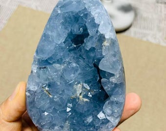 May include: A large, polished celestite crystal geode in shades of light blue. The crystal has a rounded, irregular shape with a hollow center filled with crystalline structures. The geode is held in a hand, showcasing its size and texture.