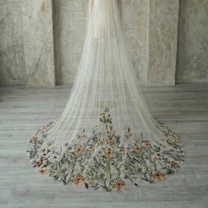 May include: A long, ivory wedding veil with a floral embroidered border. The veil is displayed on a mannequin, showcasing the delicate fabric and intricate flower design. The flowers are in shades of orange, yellow, and green.