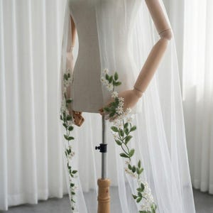 May include: A sheer white bridal veil with delicate floral embroidery. The veil features a cascade of embroidered white flowers and green leaves along the edges. The veil is displayed on a mannequin, showcasing its flowing design.