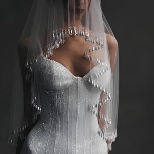 May include: A white bridal gown and veil. The dress features vertical rows of tiny beads and a sweetheart neckline. The sheer veil is decorated with hanging, teardrop-shaped embellishments. The overall style is elegant and suitable for a wedding.