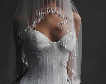 May include: A white wedding dress and veil. The dress is embellished with vertical rows of small beads and has a sweetheart neckline. The veil is sheer and adorned with dangling teardrop-shaped embellishments. The overall style is elegant and bridal.