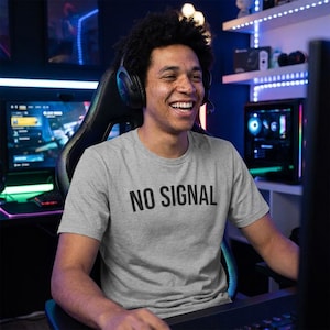 Peut inclure: Un t-shirt gris clair avec l'inscription "NO SIGNAL" en noir. La personne porte un casque et sourit. L'arrière-plan comprend des écrans d'ordinateur et du matériel de jeu.