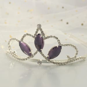 Puede incluir: Una tiara plateada con un diseño delicado presenta tres grandes piedras de imitación ovaladas de color púrpura. La tiara está adornada con piedras de imitación más pequeñas y transparentes.