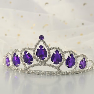 Peut inclure: Une tiare argentée avec des strass en forme de goutte d'eau violette. La tiare est décorée de plus petits strass transparents.