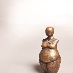 May include: Bronze-colored sculpture of a stylized female figure with crossed arms and a rounded abdomen. The sculpture has a smooth, metallic finish and a minimalist design, suitable for home decor.