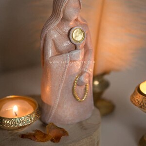 May include: A pink statue of a woman holding a mirror with a gold-colored face. The statue is wearing a draped garment and a gold-colored necklace. The statue is on a wooden base, with a lit candle in a gold-colored holder.