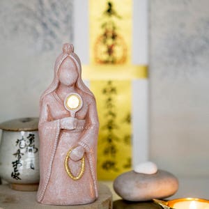 May include: A pink statue of a robed figure holding a circular object, possibly a mirror. The statue has a gold-colored ring and is on a wooden base. A small candle holder and a stack of stones are in the foreground.