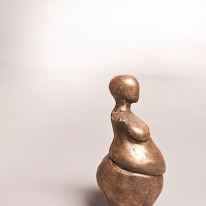 May include: A bronze-colored sculpture of a stylized human figure. The figure has a rounded base, a midsection with a curved form, and a slender neck leading to a small head. The sculpture has a textured surface.