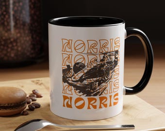 May include: A white ceramic mug with a black rim and handle. The mug features a black and white illustration of a race car with the word "NORRIS" repeated in orange text.