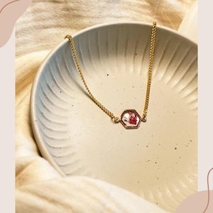 Pode incluir: Um colar dourado com um pingente hexagonal contendo flores vermelhas secas. A corrente fina e o pingente se destacam em um fundo claro, criando uma estética minimalista.