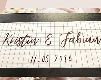 May include: A rectangular white adhesive transfer sheet featuring a black grid pattern. The text Kristin & Fabian is printed in a dark brown cursive script, with the date 11.05.2014 printed below in a simple, clean, sans-serif black font.