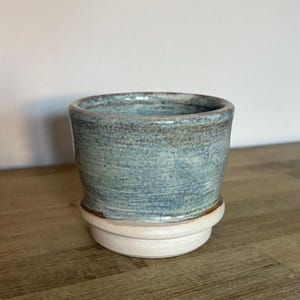 May include: A handcrafted ceramic tumbler with a light blue and brown textured glaze. The cup sits on a white base and has a slightly flared rim. The pottery is displayed on a wooden surface.