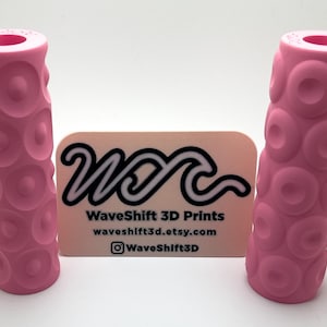 May include: Two pink 3D printed cylindrical handles with a textured surface. The handles are made by WaveShift 3D Prints, waveshift3d.etsy.com, @WaveShift3D.