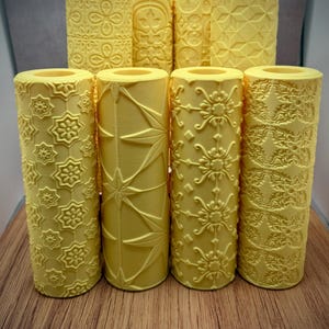 May include: A collection of yellow, patterned rolling pins. The rolling pins feature various embossed designs, including floral, geometric, and starburst patterns. The rolling pins are cylindrical and stand upright on a wooden surface.