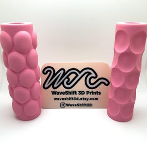 Può includere: Due forme cilindriche testurizzate stampate in 3D di colore rosa. I cilindri presentano un motivo di cerchi in rilievo. Il testo "WaveShift 3D Prints" è su un biglietto da visita sullo sfondo.