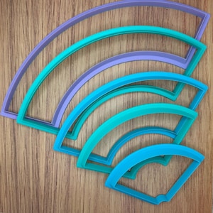 May include: A collection of five colorful plastic rainbow-shaped cookie cutters in shades of purple, teal, and blue. The cutters are arranged in a nested pattern on a wooden surface, showcasing their graduated sizes.