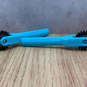 May include: Two turquoise Gratemate tools with black, gear-shaped cutting wheels. The handles are inscribed with the words "WAVESMITH" and "GRATEMATE". The tools are designed for cutting or grating. The tools are on a wooden surface.