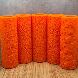 May include: Five orange textured rollers with autumn-themed designs. The patterns include pumpkins, leaves, and a spider web. The rollers are cylindrical and made of a solid orange material, perfect for crafting or decorating projects.
