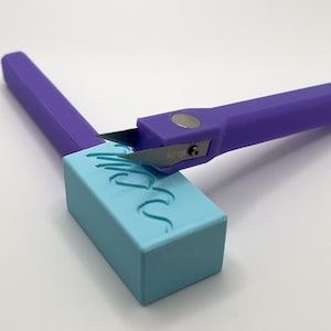 Puede incluir: Un cuchillo artesanal morado con una hoja plateada está cortando un bloque rectangular azul claro. El bloque tiene un diseño ondulado tallado.