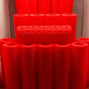 Op de afbeelding: Een verzameling helderrode cilindrische rollers met diverse textuurpatronen. De rollers zijn verticaal gerangschikt en tonen verschillende ontwerpen, waaronder geometrische en bloemmotieven. De afbeelding benadrukt de verscheidenheid aan texturen en vormen.