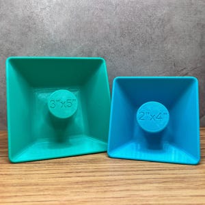 Peut inclure: Deux contenants en plastique de forme carrée avec un centre circulaire surélevé. Le plus grand récipient est vert sarcelle et mesure 3" x 5". Le plus petit récipient est bleu et mesure 2" x 4".