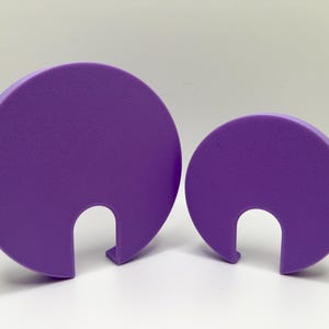 Puede incluir: Dos tapas de plástico morado con un asa recortada. La tapa más grande está en el fondo y la más pequeña está en primer plano.