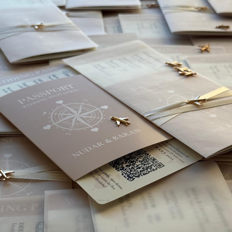 May include: Light beige passport-style wedding invitations with gold airplane charms. The invitations feature a compass rose design and the words "Passport Wedding Invitation". The names "NUDAR & BARAN" are printed on the front of the invitation.