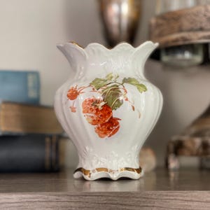 May include: White ceramic vase with a scalloped rim and gold trim. The vase features a hand-painted design of red strawberries with green leaves. A decorative piece for home decor.