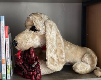 May include: A plush, cream-colored dog toy with long, floppy ears and a red collar. A dark red decorative object is in front of the dog. Books with titles like "The Mindfulness Workbook for OCD" are visible on a shelf.