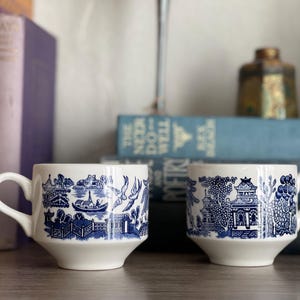May include: Two white ceramic mugs with blue Willow pattern designs. The mugs have a classic shape with a handle and a slightly flared base. The background includes books and decorative items.