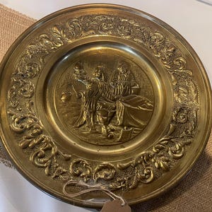 May include: A brass wall plate with an ornate design featuring two figures in relief. The plate has a raised rim and a smooth, polished surface.