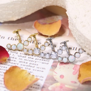 Può includere: Tre orecchini a polsino curvi. Due sono dorati, uno con pietre simili all'opale e l'altro con cristalli trasparenti. Il terzo è argentato con cristalli trasparenti e pietre simili all'opale. Gli orecchini sono su carta.