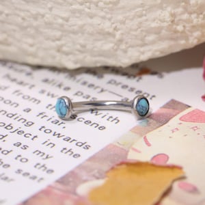 May include: A curved eyebrow piercing with a silver bar and turquoise stone accents. The jewellery is displayed on a page with printed text and a decorative background.