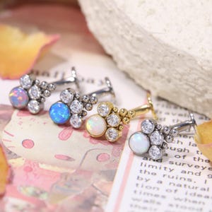 May include: Four pieces of jewelry with a cluster design. Each piece features a central gemstone, surrounded by smaller clear stones. The gemstones are opal, blue, and clear. The posts are silver and gold.