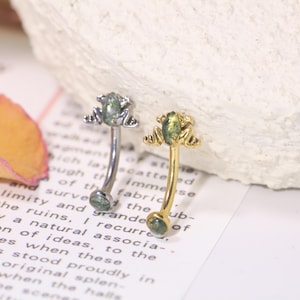 May include: Two curved eyebrow rings. One is silver-toned, the other gold-toned. Each features a small frog charm with green accents. The rings are displayed on a vintage book page.