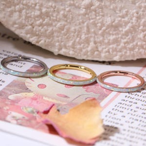 May include: Three thin rings with an opal-like inlay. One ring is silver, one is gold, and the third is rose gold. The rings are displayed on a pink and white surface with text.
