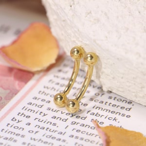May include: A pair of gold-coloured, curved barbell earrings. Each earring has a small ball at each end. The earrings are displayed on a white surface with text from a book visible in the background.