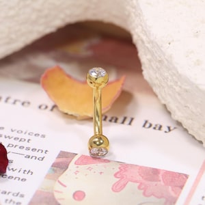 Puede incluir: Un piercing curvo de color dorado con una gema redonda y transparente en cada extremo. La joya se muestra sobre un fondo rosa y blanco con texto.