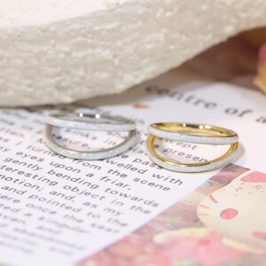 May include: Two rings with a double-band design. One ring is silver-toned, the other gold-toned. Both rings feature a white opal-like inlay. The rings are displayed on a printed paper surface.