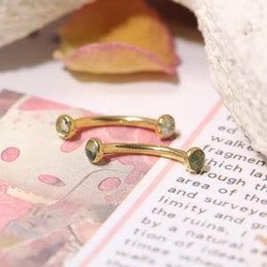 May include: Two curved, gold-coloured eyebrow rings, each with small, round, light green and white gemstones at the ends. The jewellery is displayed on a patterned surface with text and a blurred background.