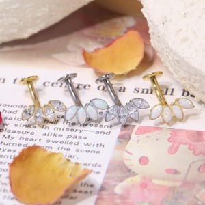 May include: Three belly button rings with a floral design. One is gold-toned with clear crystals, another is silver-toned with white opal-like stones, and the third is gold-toned with white opal-like stones. The jewelry is displayed on a printed surface.