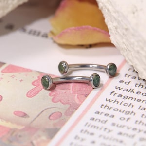Puede incluir: Dos piercings curvos con forma de barra, con metal plateado y extremos de piedra verde. Las joyas se exhiben sobre una superficie estampada con texto y un pétalo de rosa en el fondo. Los piercings son para modificación corporal.