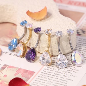 May include: A collection of belly button rings with teardrop-shaped gemstones in various colors, including blue, purple, and clear. The rings have curved, silver-colored bars and are displayed on a textured surface.