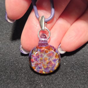 Close-up of a colorful heady glass pendant with a dainty frit implosion design in shades of violet, magenta, and amber, shown against a black background.