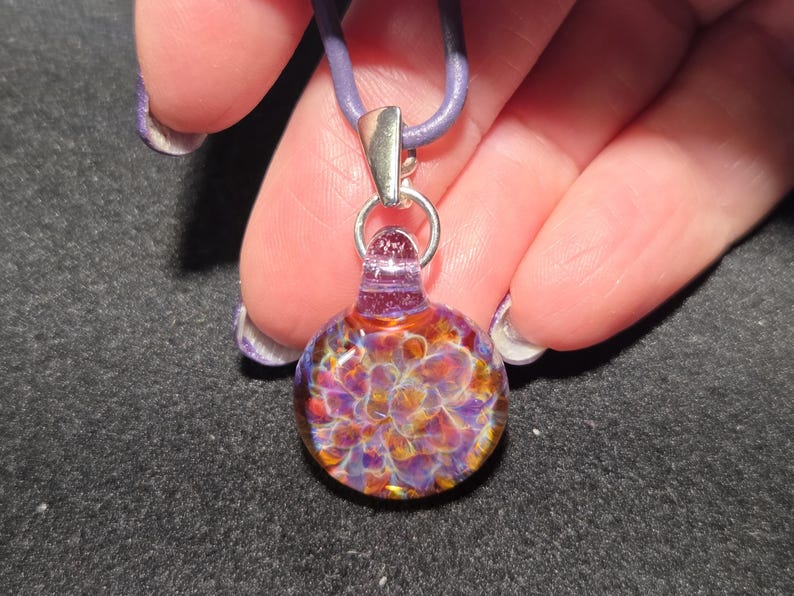 Close-up of a colorful heady glass pendant with a dainty frit implosion design in shades of violet, magenta, and amber, shown against a black background.